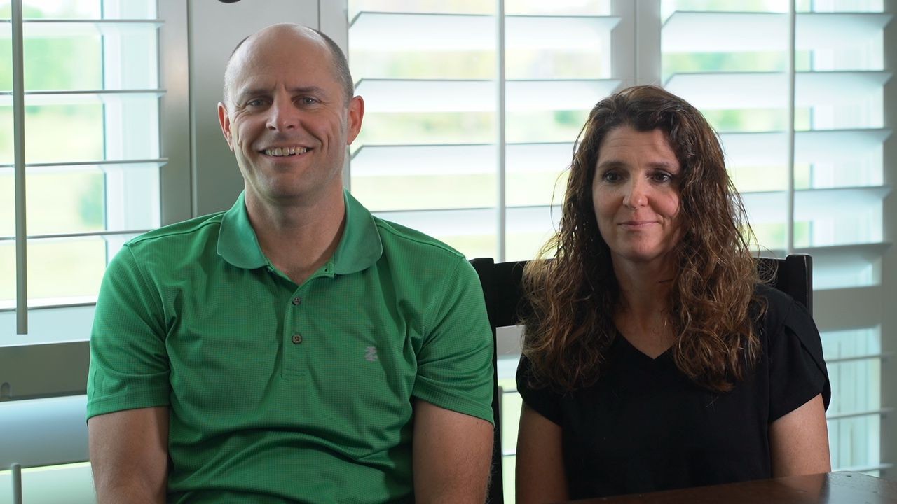 A pest control client with light skin is seated on the left, smiling directly at the camera. He is wearing a green collared polo shirt. To his right, a woman with long, curly brown hair and light skin is seated, looking towards the camera with a slight, gentle smile. She is wearing a black V-neck top. They appear to be indoors, possibly at a table, with a large window featuring white horizontal blinds in the background.