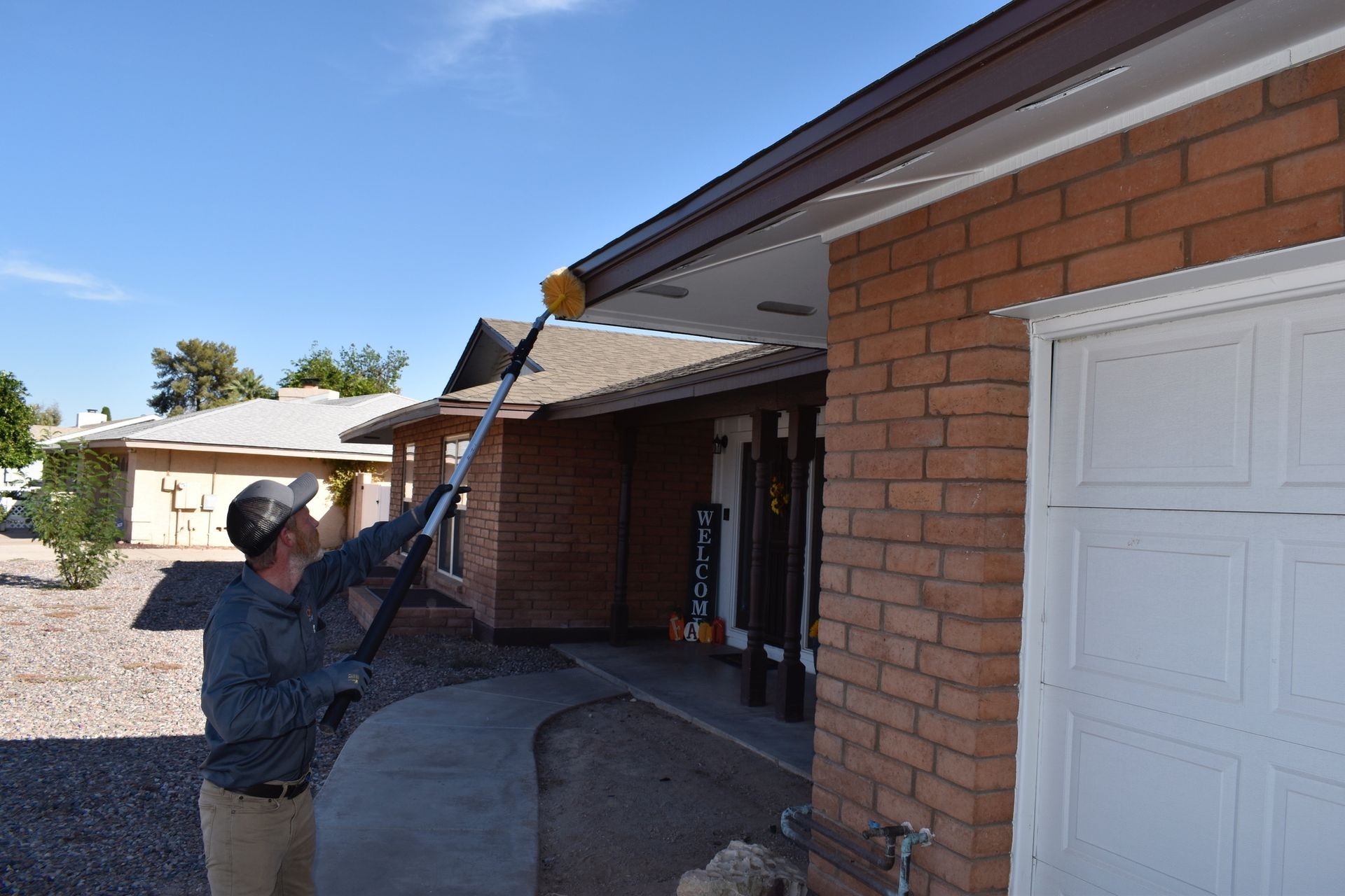 A pest control technician, seen from the waist up, stands outdoors, facing a brick house with a white garage door to the right. He is wearing a gray long-sleeved shirt, tan pants, and a gray baseball cap with a dark brim. He is holding a long-handled duster with a fluffy yellow head, extending it upwards towards the eave of the house. The sky is blue with minimal clouds. In the background, other houses and landscaping are visible.
