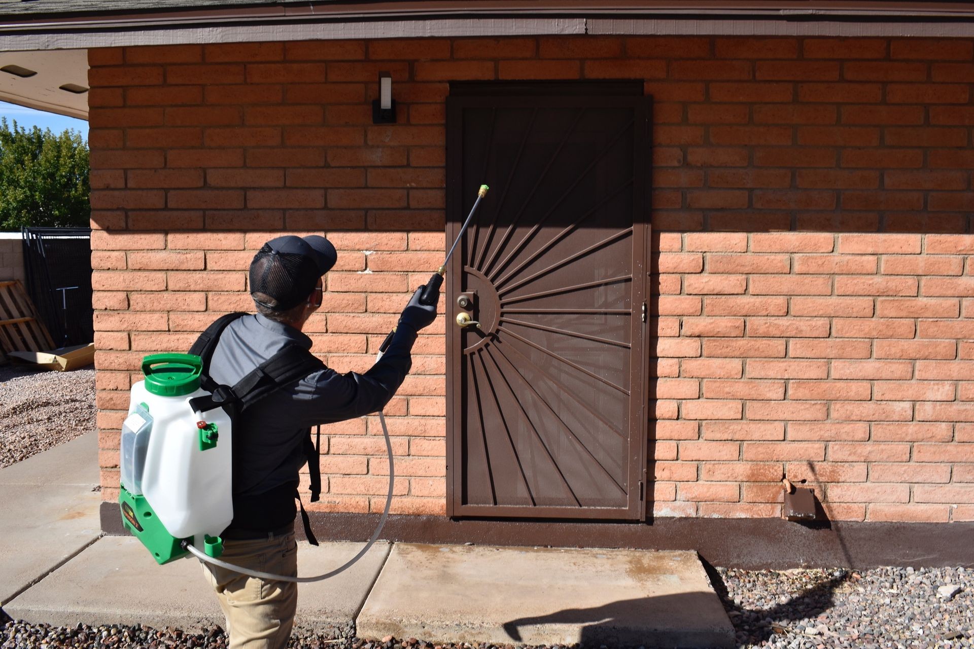 A pest control technician, seen from behind, stands outdoors, facing a brick house with a dark brown metal security door. The technician is wearing a dark gray long-sleeved uniform shirt, light-colored pants, gloves, and a dark baseball cap. He is holding a white and green backpack sprayer and is applying a spray with a wand directed towards the base of the house and the door. The ground is concrete, and there's a gravelly area to the left. The sun casts strong shadows.